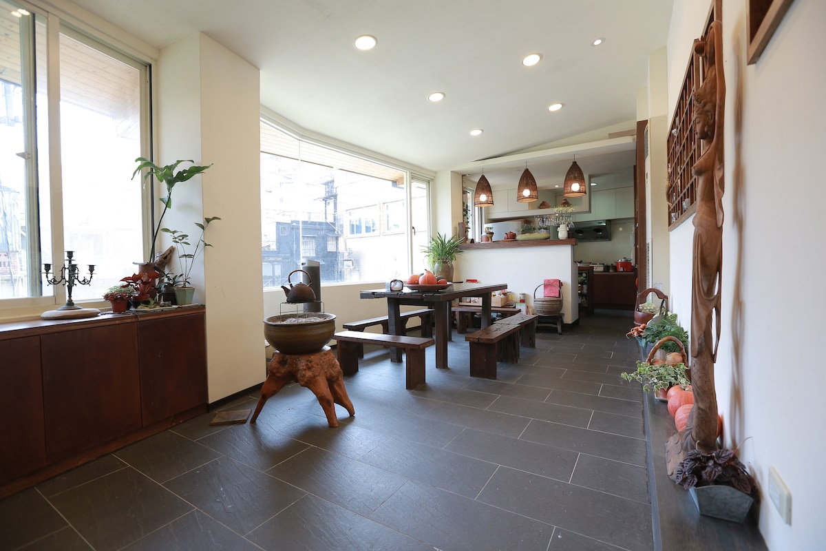 A spacious living area is depicted, featuring large windows that allow natural light to fill the room. A wooden dining table is centrally located, surrounded by benches. Decorative elements, like plants and rustic art pieces, add character to the inviting interior.