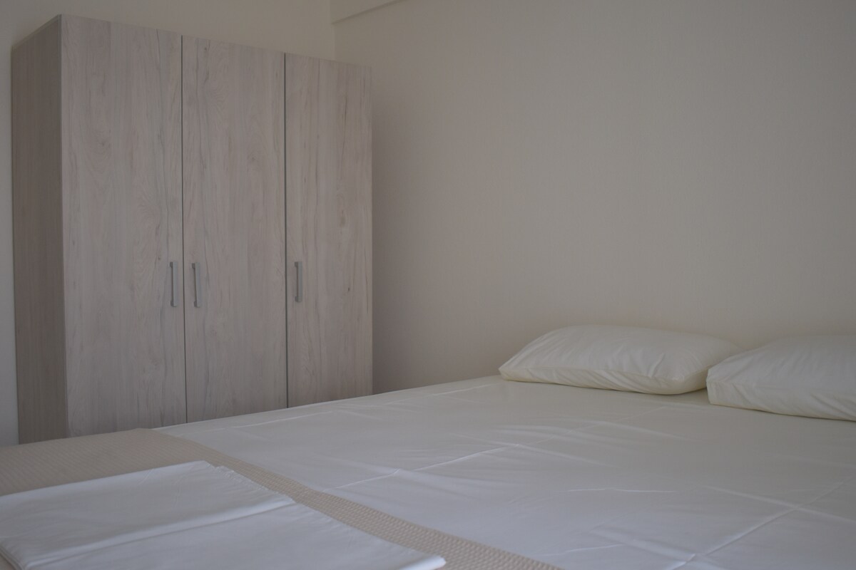 A spacious sleeping area features a light-colored bed with two soft pillows. A wooden wardrobe with a natural finish stands beside the bed, offering storage space. Neutral wall colors contribute to a calm atmosphere.