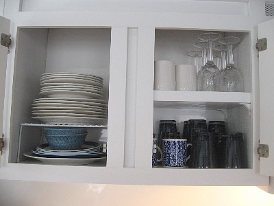A white kitchen cabinet displays neatly organized dinnerware. The upper section contains white plates and colorful bowls, while the lower section features various cups and glasses, including stacked blue and black mugs and several clear glassware items.