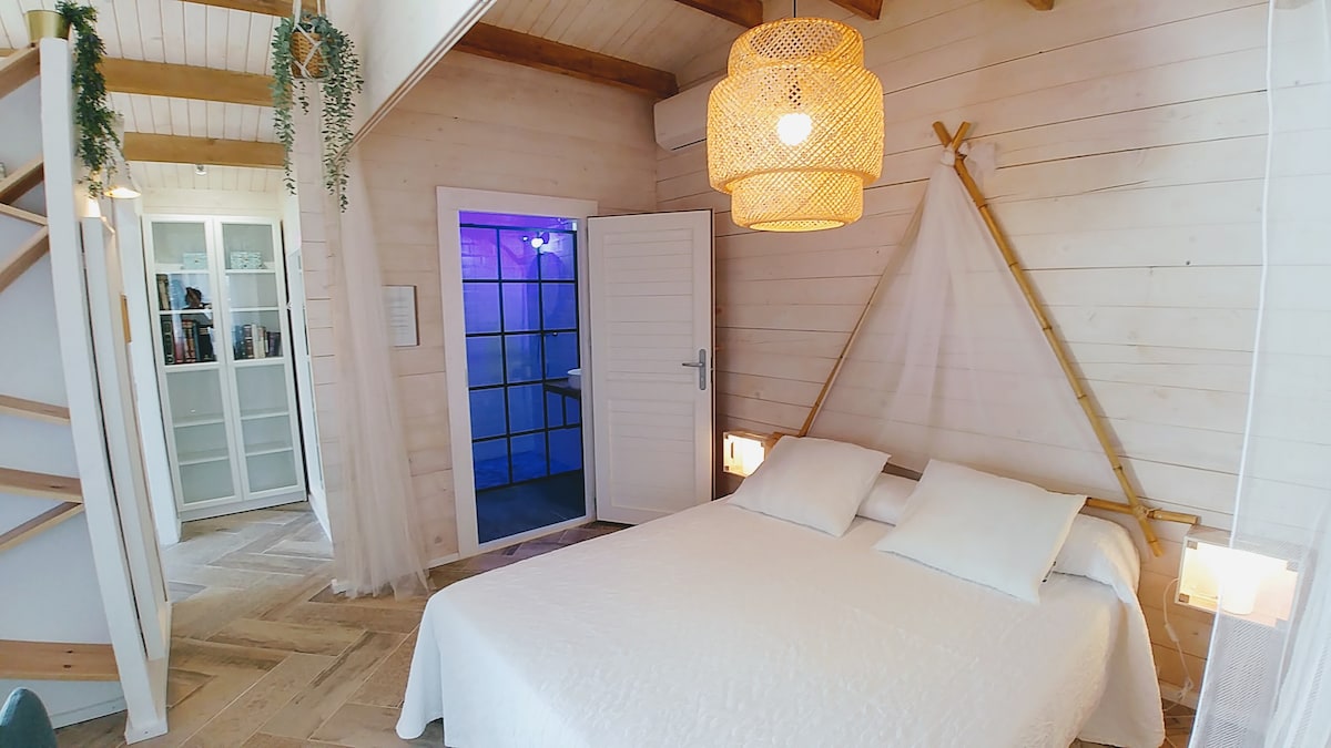 An open and airy sleeping area is present, featuring a spacious bed dressed in crisp white linens. Attention is drawn to the wooden ceiling and light fixtures. A doorway leads to another section of the cabin, while a lush plant decorates the environment.