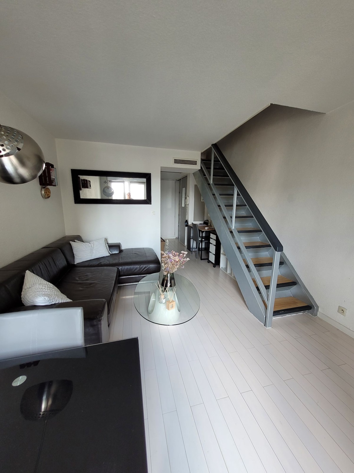 A spacious living area features a comfortable sectional sofa and a glass coffee table. A staircase leads to the upper level, while a large window allows natural light to illuminate the room. Decorative elements including a mirror enhance the space's ambiance.