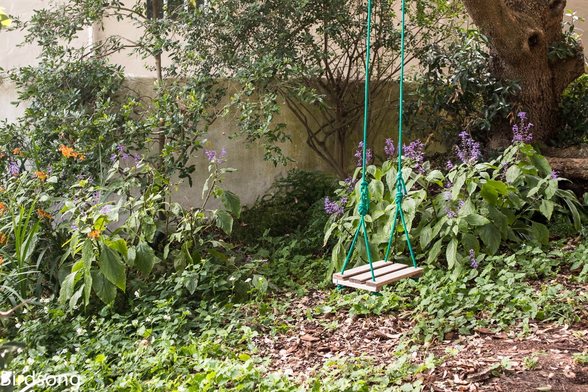 A wooden swing is suspended from a sturdy tree branch, surrounded by lush greenery. Colorful flowers, including purple blooms, peek through the vibrant foliage, creating a serene and inviting outdoor space. Soft, natural light filters through the trees, enhancing the peaceful atmosphere.