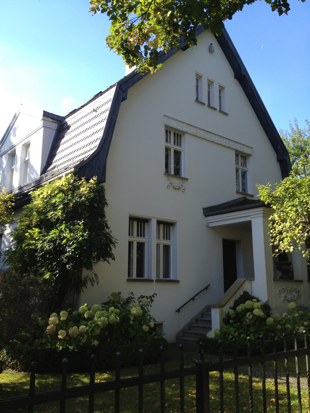 A charming house is depicted, featuring a steeply pitched roof and decorative architectural details. Well-maintained gardens with flowering plants surround the entrance, which is accessed by a set of steps. Light filters through the large windows, enhancing the welcoming facade.