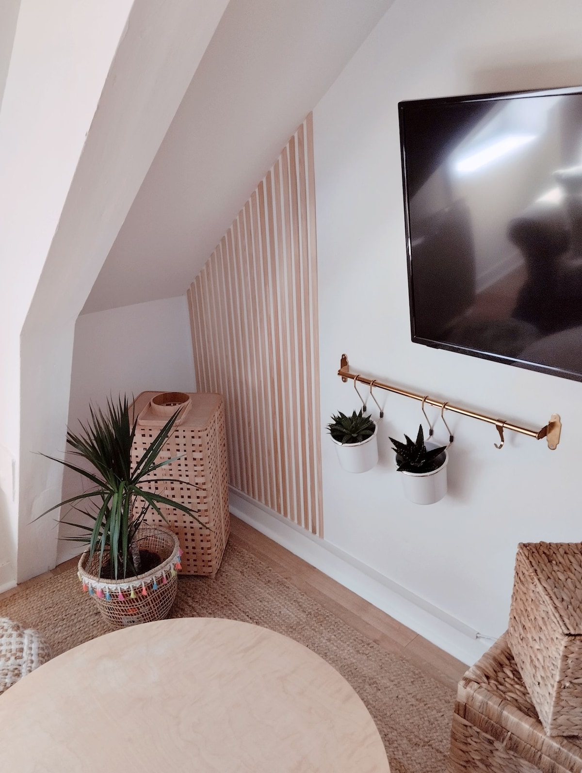 A cozy corner of the living room is highlighted by a mounted television, minimalist wall decor, and indoor plants. Natural light is allowed to filter through the space, complementing the subtle textures of the woven area rug and cabinetry.