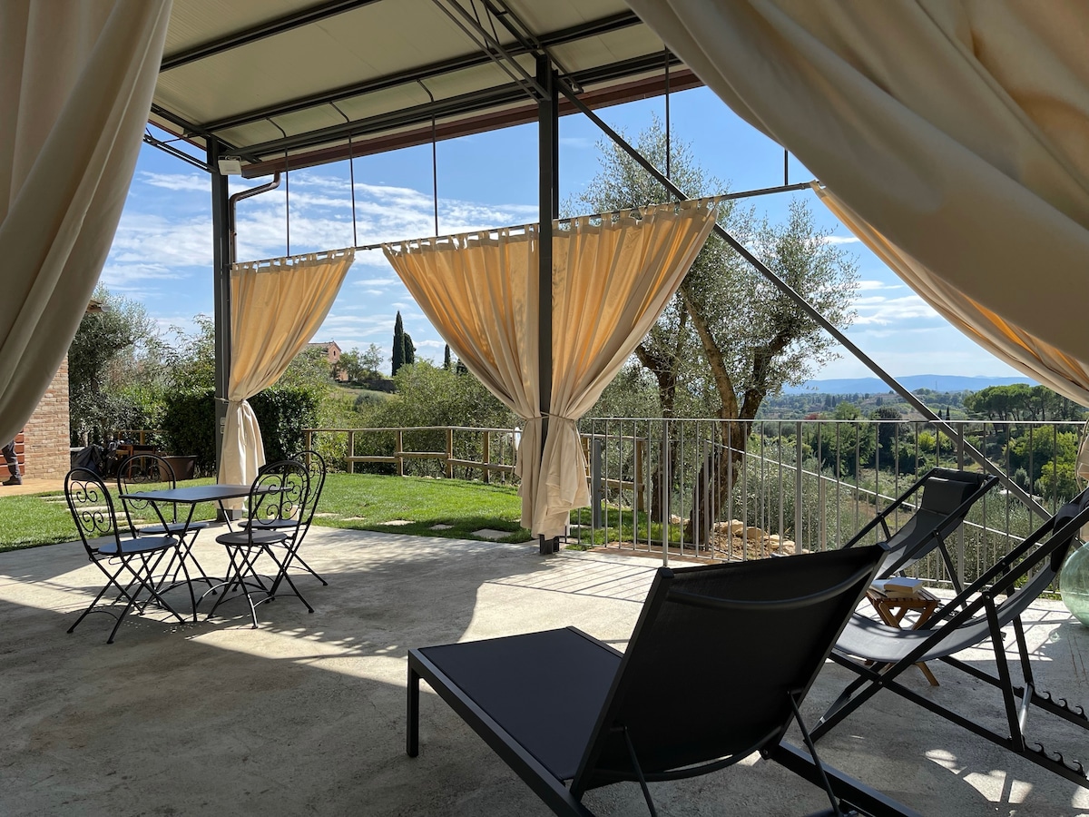An outdoor seating area features a small round table with two chairs and a lounge chair under a shaded terrace. Curtains gently frame the space, while olive trees and greenery extend into the background, offering views of the surrounding landscape.