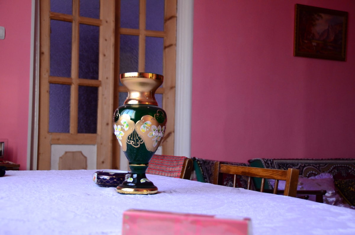 A decorative green vase with gold accents is placed at the center of a white table. The background features walls painted in a soft pink hue, with wooden doors partially visible, and patterned textiles draped over nearby seating.