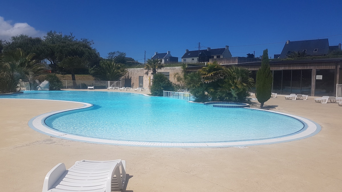 The outdoor swimming pool features a large, curved shape with inviting turquoise water. Surrounded by palm trees and greenery, the area is sunlit and spacious, with several lounge chairs positioned around the pool deck, creating an ideal space for relaxation.