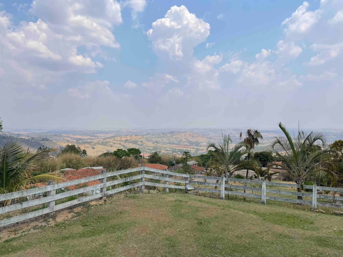 A serene view showcases rolling hills and a vast landscape under a cloudy sky. A white wooden fence encloses the grassy area, framing the expansive scenery with patches of green and hints of distant structures blending into the horizon.