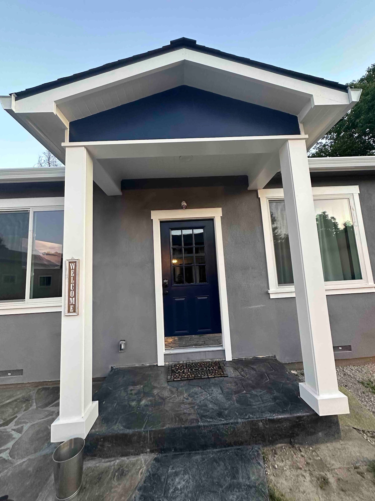 The entrance of the home features a navy blue door framed by white columns. A welcome sign is mounted on the side, with a stone pathway leading to a stone porch. Large windows beside the door allow for natural light.