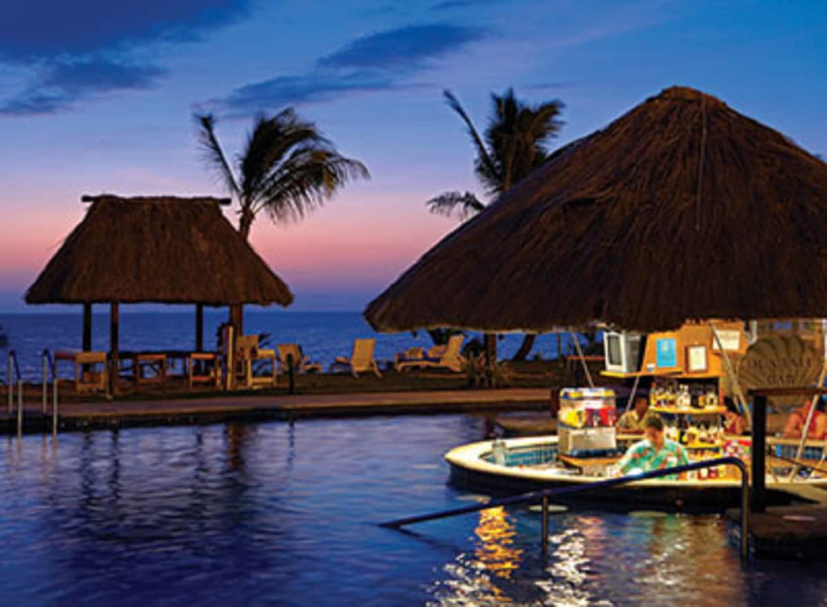 A swim-up bar is visible in the foreground, surrounded by calm water. Two thatched-roof structures can be seen in the background, set against a colorful twilight sky and gentle ocean waves. Lounge chairs line the pool area, enhancing the resort's relaxed atmosphere.