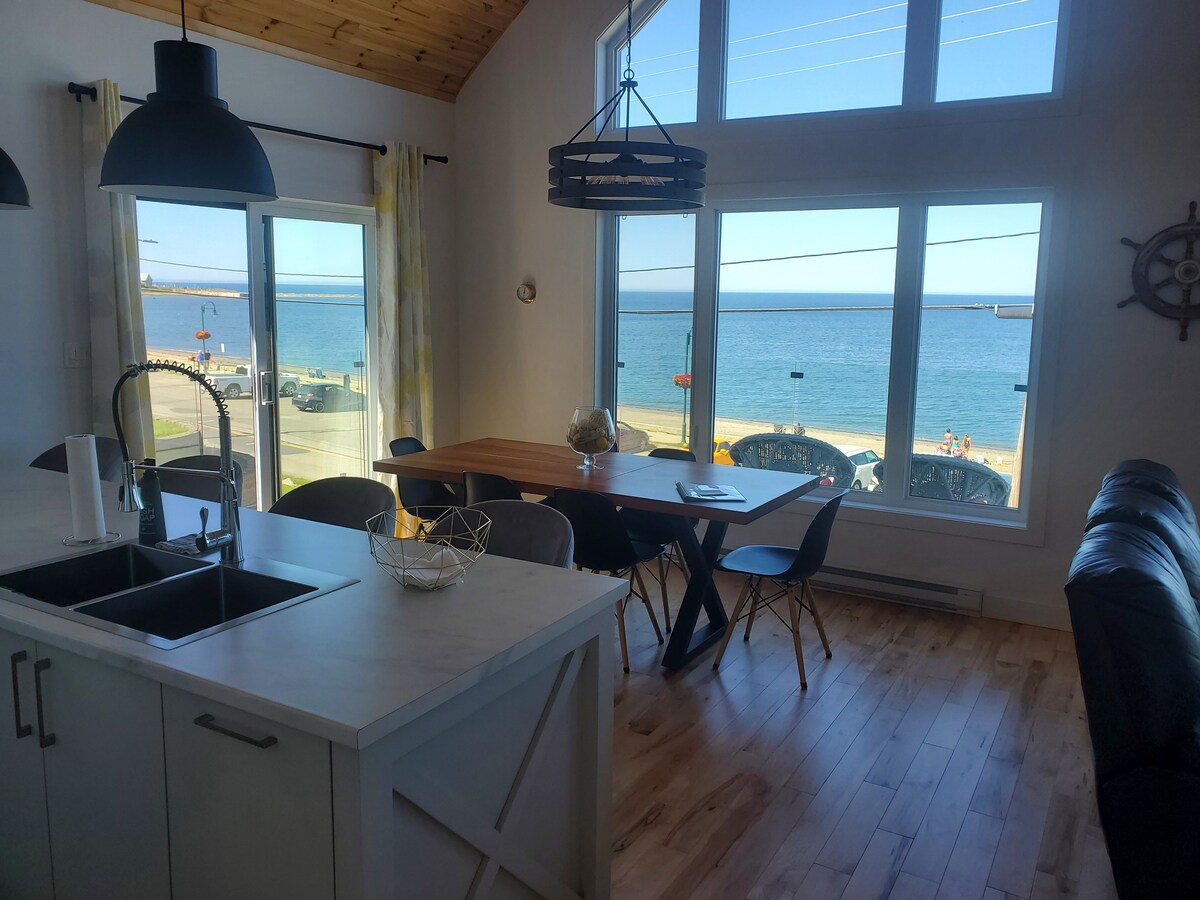 A bright dining area features a large wooden table surrounded by chairs, with a view overlooking the sea through expansive windows. The kitchen is designed with a modern aesthetic, including a sleek countertop and pendant lighting, creating an open and inviting space.