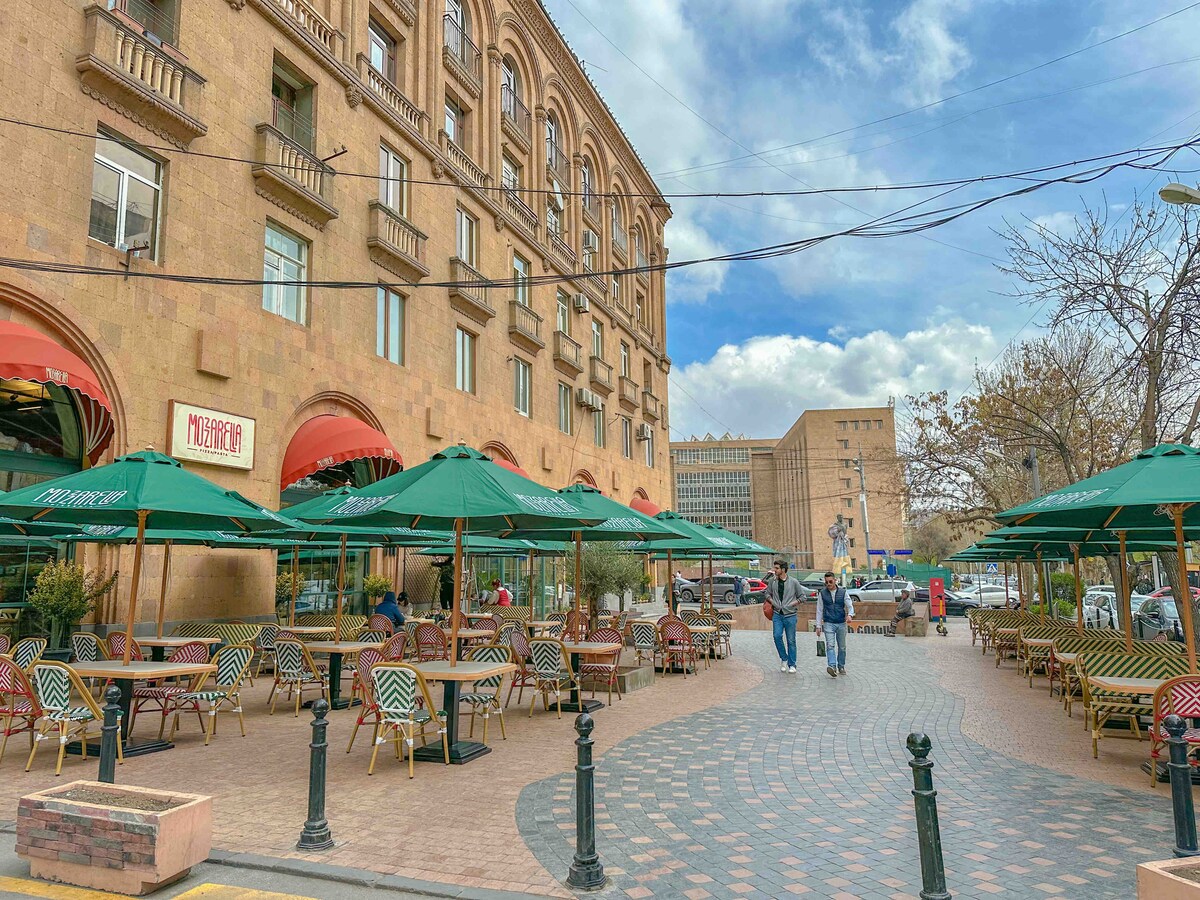 An outdoor dining area is arranged with green umbrellas and wicker furniture, situated adjacent to a historic building. Guests are seen enjoying their time at the tables. The surrounding atmosphere features a combination of cobblestone pavement and trees, creating a welcoming environment.