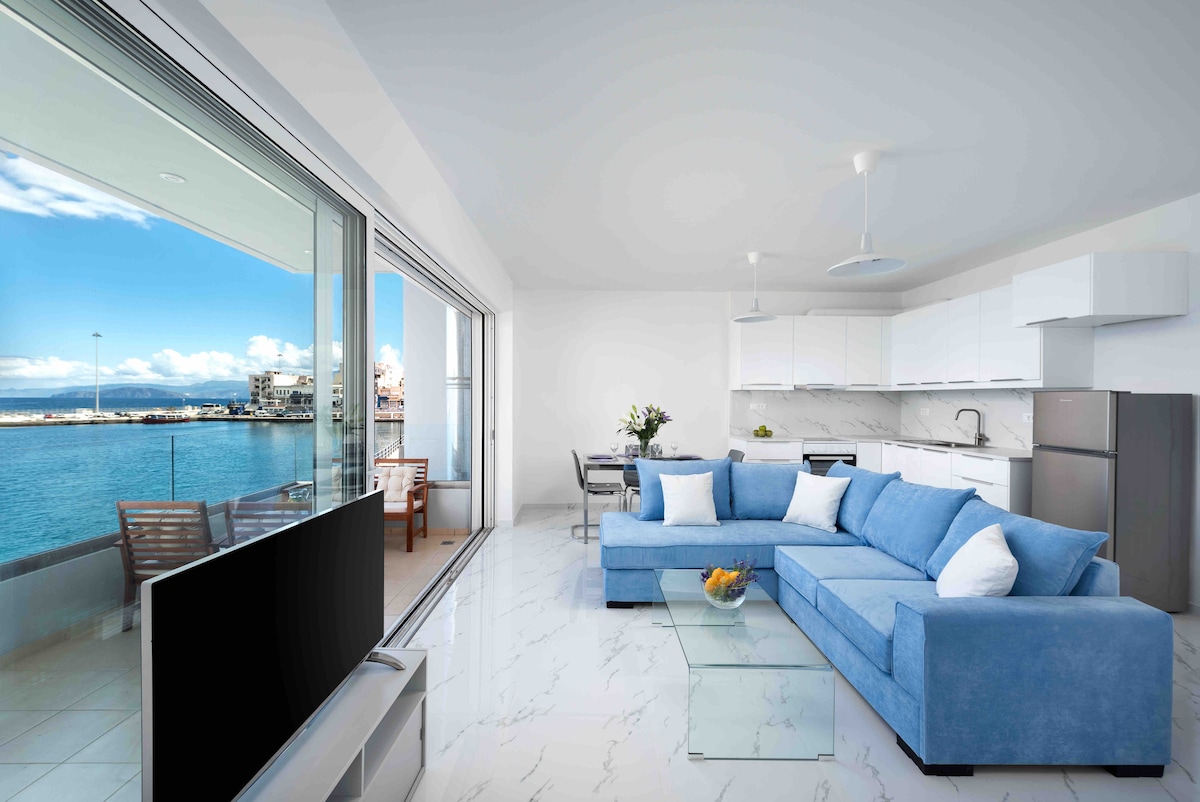 A modern living area features a blue sectional sofa and a glass coffee table, with expansive windows offering views of the sea and scenic harbor. A dining area is visible, alongside a contemporary kitchen equipped with white cabinetry and stainless steel appliances.