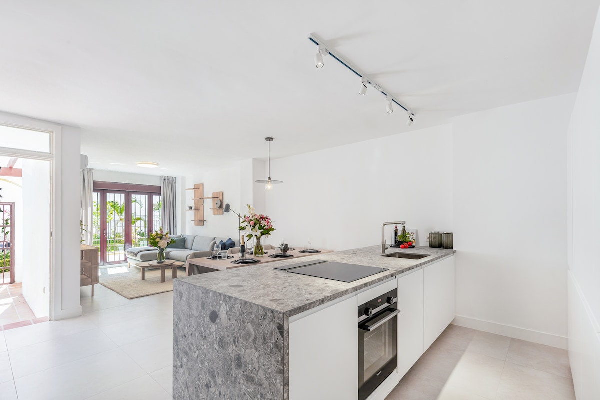 An open-concept kitchen showcases a spacious granite countertop with a modern stove and built-in oven. Natural light fills the area, illuminating the sleek cabinetry. Beyond, a living area can be seen with comfortable seating and access to a terrace adorned with greenery.