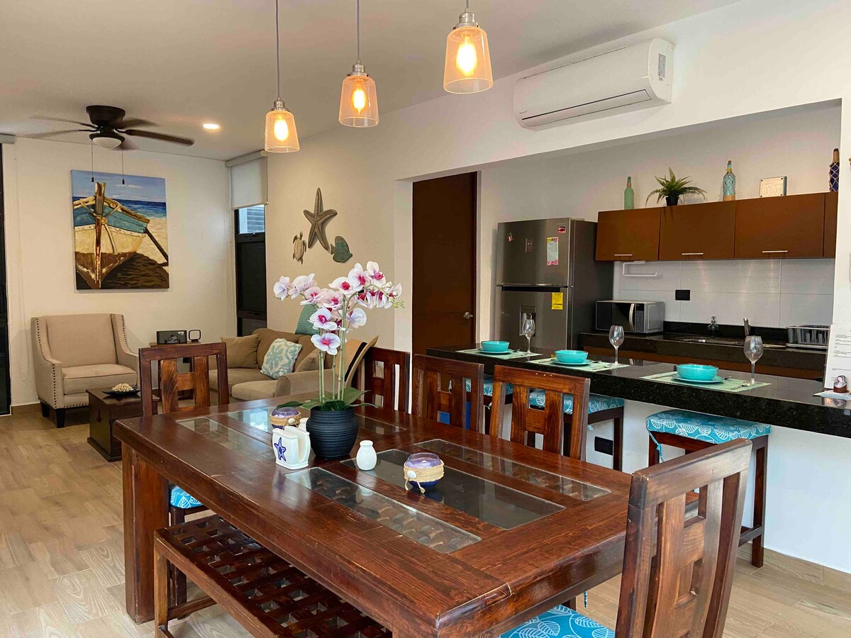 The dining area features a large wooden table surrounded by eight chairs, complemented by decorative blue cushions. Above, pendant lights illuminate the space. A modern kitchen is visible in the background, showcasing dark cabinetry and stainless steel appliances. A cozy seating area is located to the left.