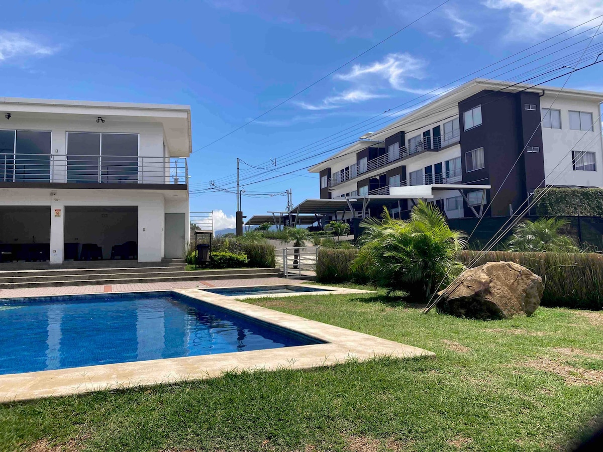 A pool area is framed by lush green grass and palm plants. Sunlight reflects off the blue water. Two modern multi-story buildings are positioned in the background, showcasing a mix of white and dark exteriors. The peaceful surroundings suggest a tranquil atmosphere.