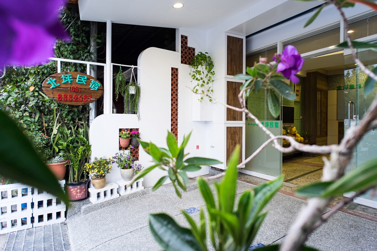 A welcoming entrance is highlighted by a blend of modern and natural elements. Potted plants with colorful flowers frame the entryway, while a sign displaying the property name is mounted on a stylish white wall. The design promotes an inviting atmosphere for guests.