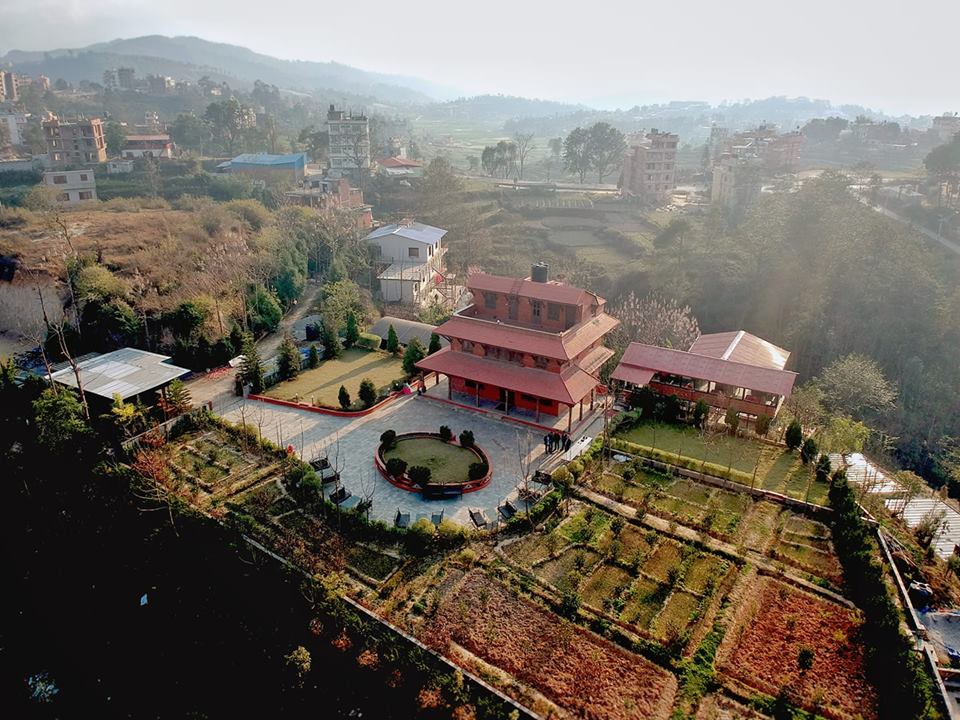 An aerial view showcases a traditional Newari home with three and a half stories, surrounded by lush gardens. The clear layout features well-maintained pathways and manicured greenery, set against a backdrop of rolling hills and distant mountains.