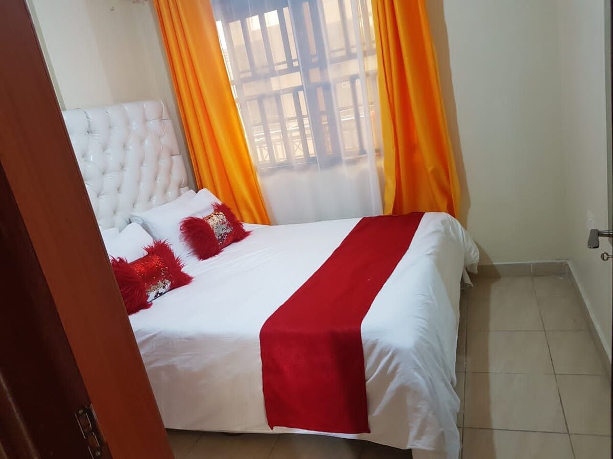 A cozy bedroom features a spacious bed dressed in white linens, accented by a red coverlet and decorative pillows. Large windows with sheer curtains allow natural light to brighten the room, complemented by vibrant orange drapes framing the view.