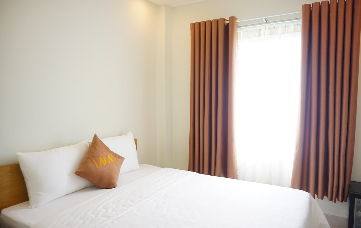 A cozy sleeping area is highlighted by a spacious bed adorned with crisp white linens. An accent pillow in warm tones rests on the bed. Floor-to-ceiling curtains in a rich amber shade frame the window, allowing soft natural light to illuminate the room.