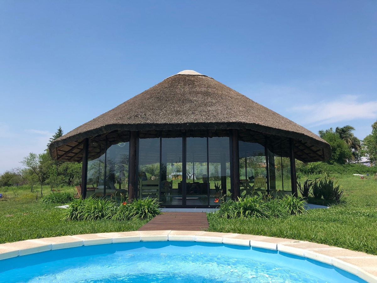 A round, thatched-roof structure is seen from the perspective of a small pool. Expansive glass walls provide a view into the interior, which is surrounded by lush green grass and plants. The tranquil outdoor environment contributes to a serene atmosphere.