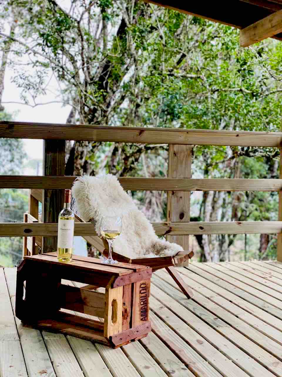 A wooden deck is shown surrounded by greenery, featuring a cozy chair draped with a soft throw. A glass of white wine and a bottle are placed on a rustic crate, inviting relaxation amidst the natural setting.