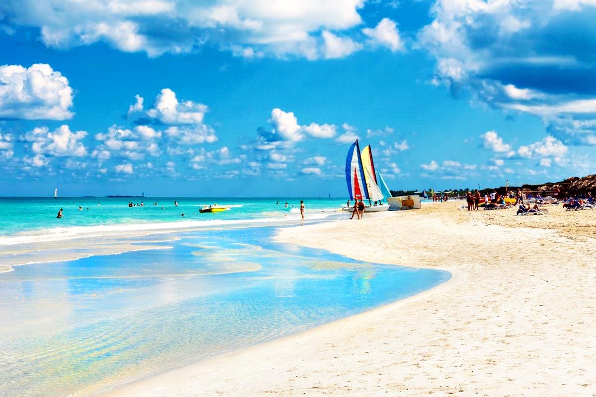 The image captures a lively beach scene featuring soft, white sand and turquoise waters. Colorful sailboats are positioned near the shore, with people enjoying various activities in the water. A clear sky adorned with fluffy white clouds enhances the vibrant atmosphere.