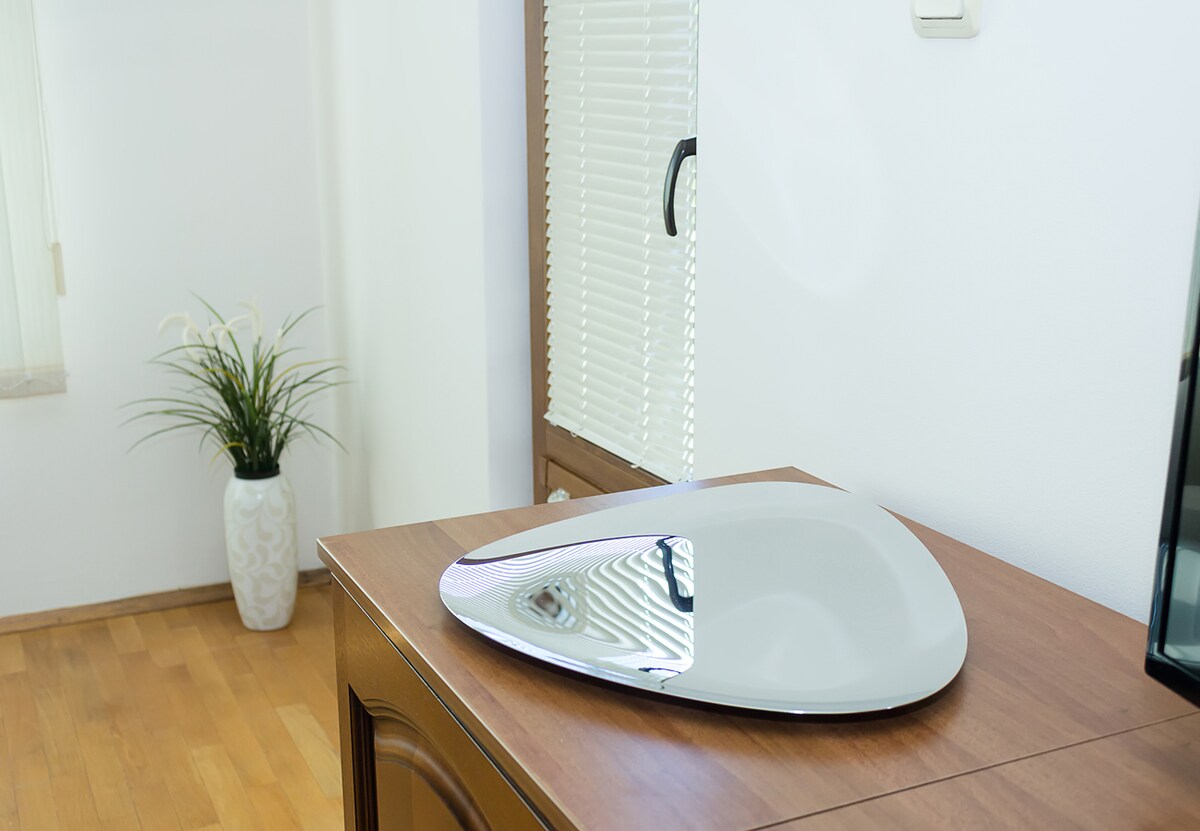 A sleek, modern decorative piece is positioned atop a wooden cabinet. Natural light reflects off its smooth surface, complemented by a potted plant in a white vase nearby. The clean lines and minimalistic design enhance the overall aesthetic of the room.