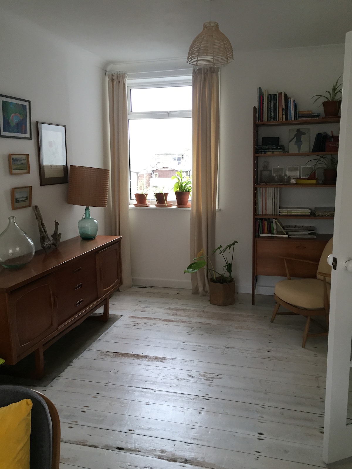 A light-filled corner of the room is presented, featuring a wooden cabinet and a vintage chair. A tall bookshelf is filled with books and decorative plants, while a lamp and a vase add subtle decor. Natural light filters through a window with sheer curtains.