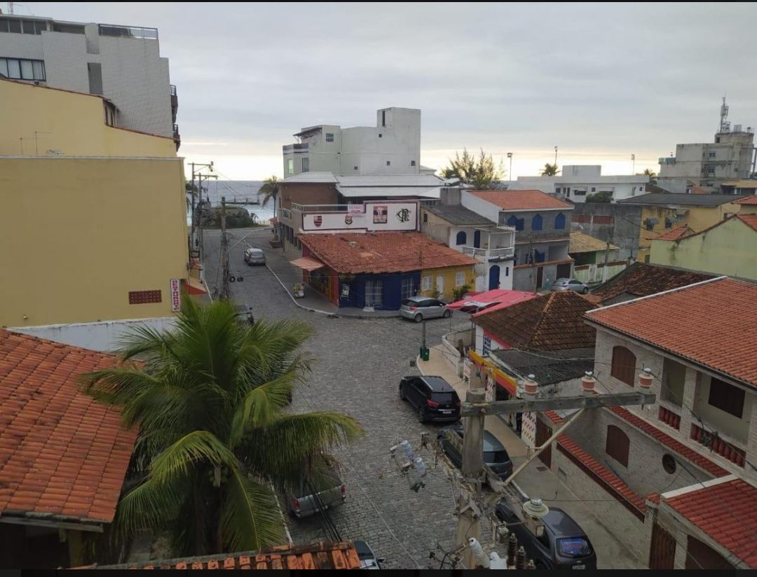 A view from above showcases a charming street with cobblestone paving, lined with colorful buildings featuring tiled roofs. Nearby, palm trees are visible, and distant structures indicate proximity to the ocean. The street is quiet, with a few parked cars and minimal pedestrian activity.