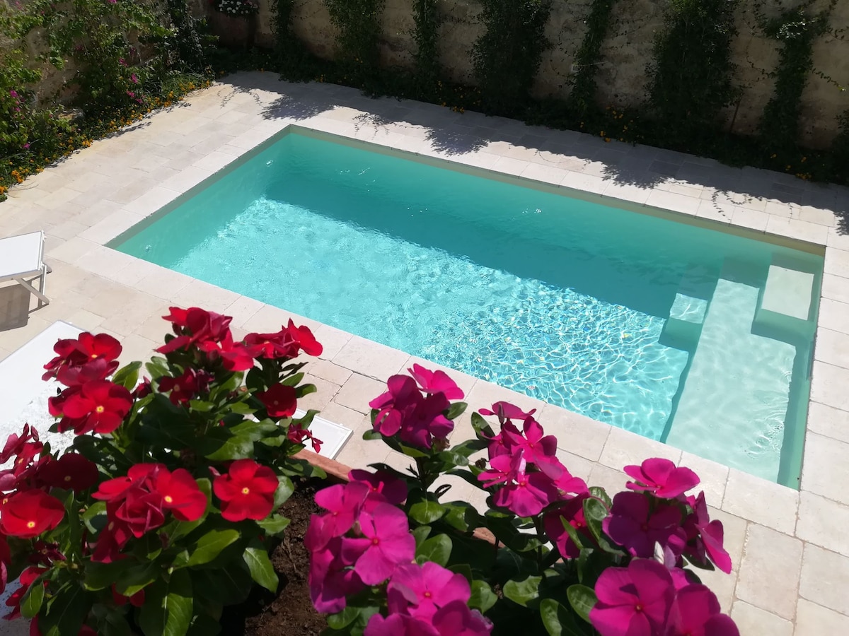 A sparkling salt-water pool is surrounded by lush greenery and vibrant pink and red flowers. The pool features a gradual entry area, providing a calming contrast against the light-colored stone patio.