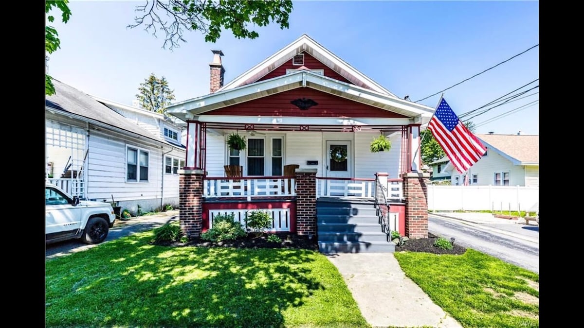 The charming bungalow features a welcoming front porch adorned with plants and two rocking chairs. A classic American flag hangs prominently, while the well-maintained lawn and flower beds add a touch of greenery. The exterior showcases traditional architectural details, creating a homely appeal.