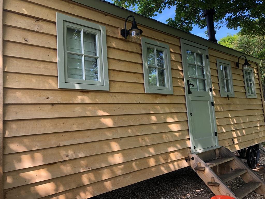 The exterior of the shepherd's hut is showcased with wooden siding in natural tones. Four windows with green frames provide a charming accent, while a light fixture hangs beside the entrance door. A small staircase leads to the inviting door, surrounded by greenery.