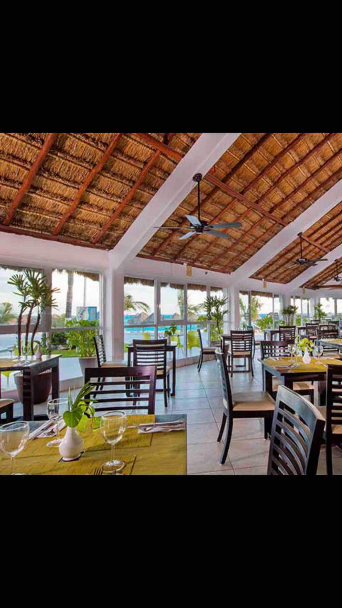 A dining area features wooden tables and chairs arranged throughout the space. Large windows provide natural light and views of palm trees and the outdoor pool. Tropical plants are placed decoratively in the room, complementing the straw-thatched ceiling.