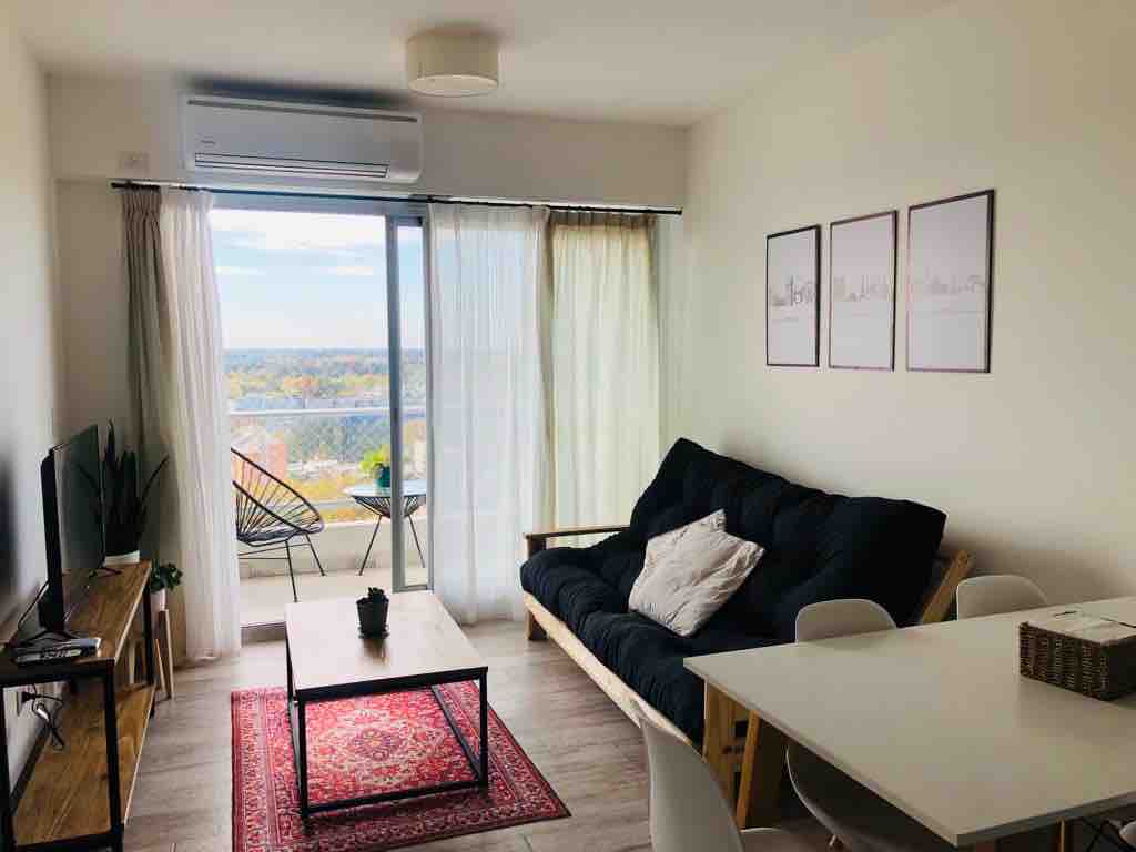A comfortable living area features a dark sofa bed, a wooden coffee table centered on a delicate area rug, and a dining table with chairs. Panoramic windows allow for abundant natural light, revealing views of the river and landscape beyond.