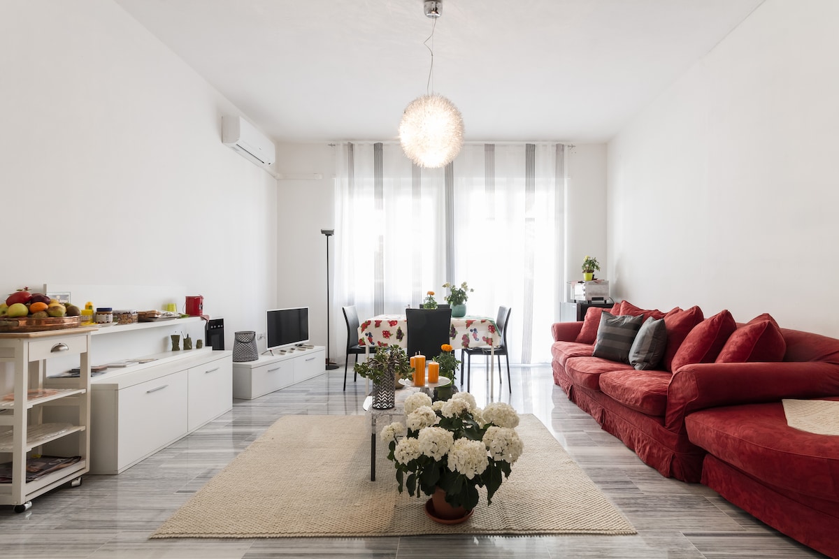 A spacious living area features a red sectional sofa and a glass dining table set for four. The room is illuminated by natural light through large windows, complemented by soft gray curtains. A decorative floral arrangement sits on a neutral rug, enhancing the space.
