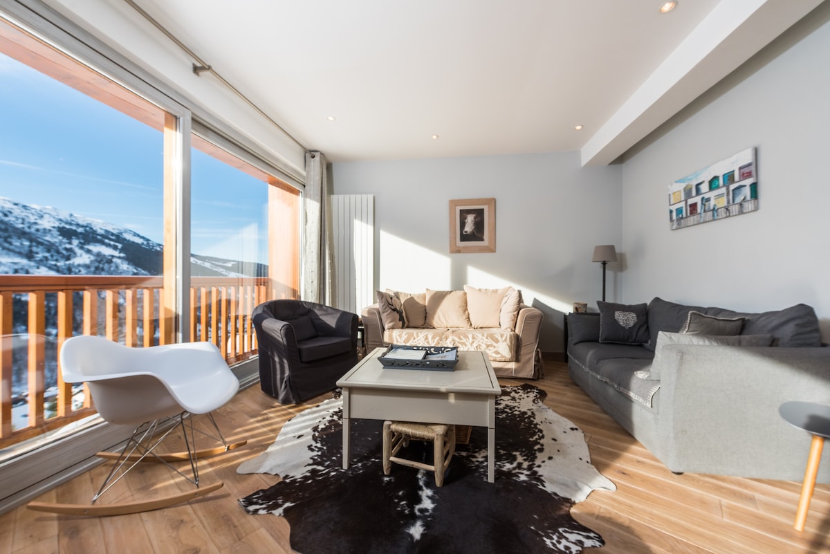 A spacious living area is highlighted by large windows that offer expansive views of the snowy valley. Comfortably arranged seating includes a light-colored sofa and dark armchairs around a central coffee table. A decorative cowhide rug adds texture to the wooden floor.