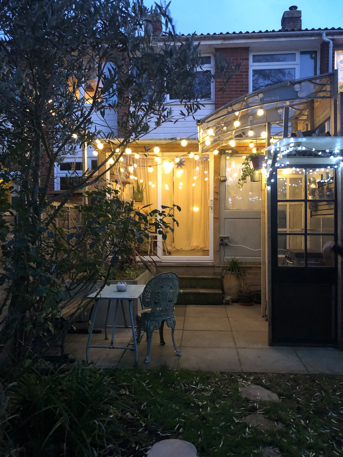A charming outdoor space features string lights overhead, creating a soft ambiance. A small table and two blue chairs are positioned on the stone patio, surrounded by greenery. A glass structure is visible on one side, offering additional space for relaxation.