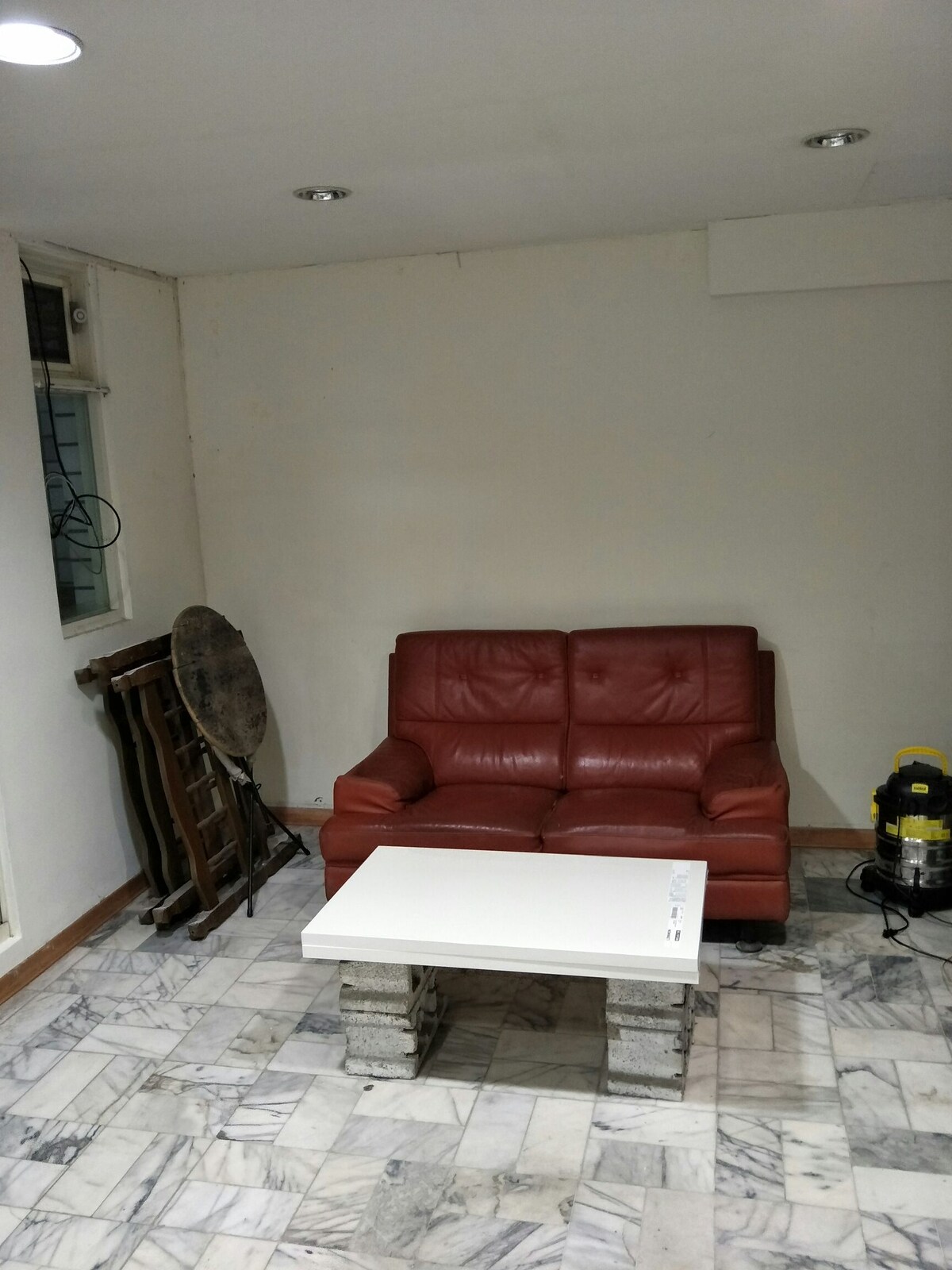 A small living area features a red leather sofa and a white coffee table made from stacked blocks. The floor is covered in light marble tiles, with a simple wall design and overhead lighting providing illumination. A window is visible on one side.