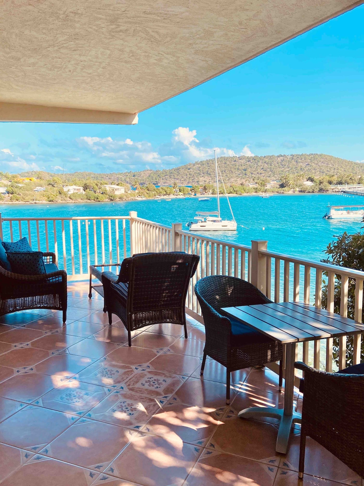 A balcony with wicker furniture overlooks the vibrant blue water. Two chairs and a small table are positioned for relaxation. The scene includes distant hills and sailboats gently bobbing in the water, creating a serene coastal ambiance.