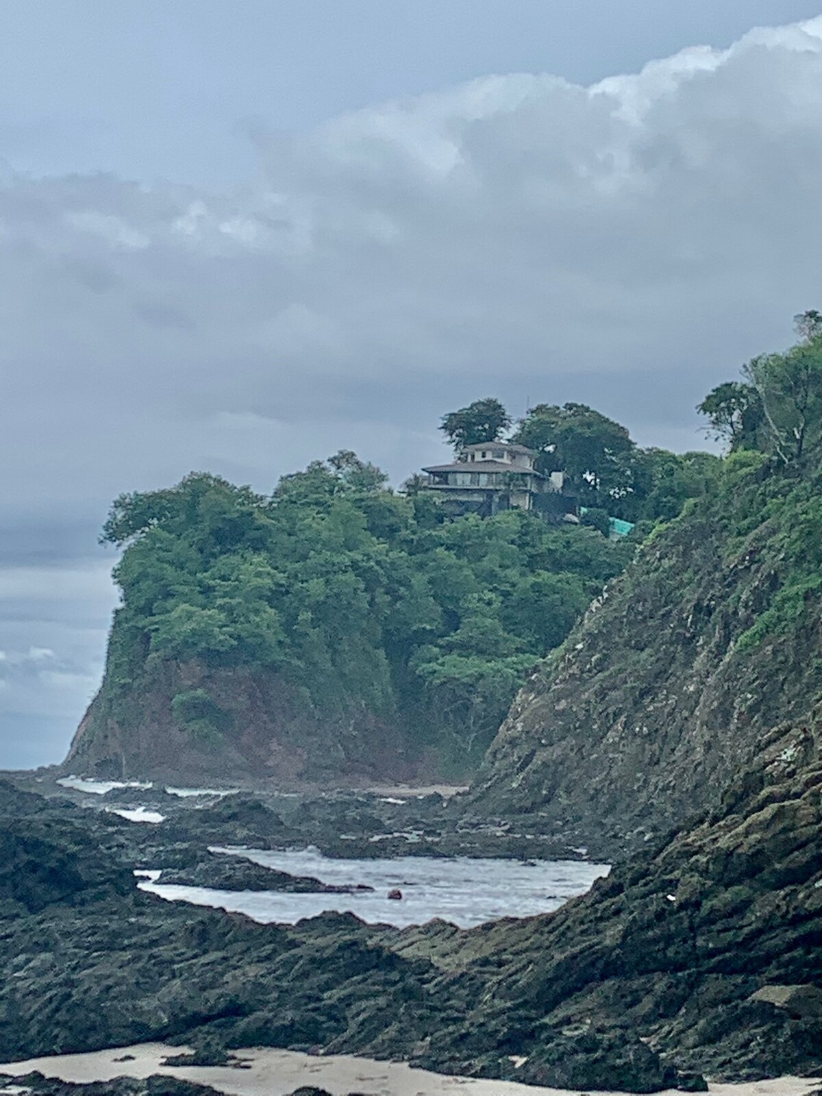 A rocky shoreline is visible in the foreground, with the ocean gently lapping against the boulders. In the distance, a lush green cliff rises, topped by a multi-level house surrounded by trees, creating a picturesque coastal scene. The sky is overcast, adding a serene ambiance.