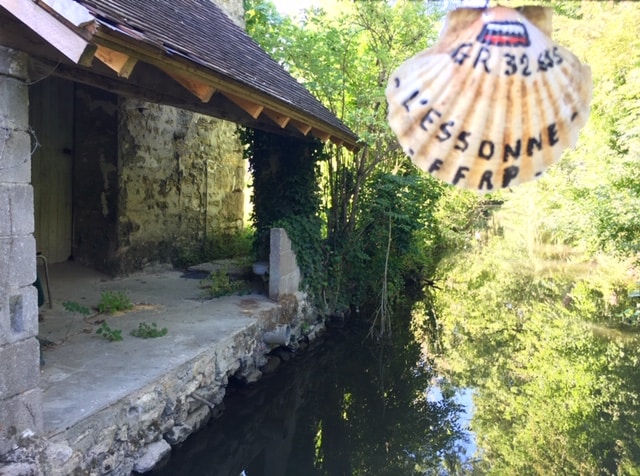 A serene view of the Essonne River, with a stone structure partially visible on the bank. Lush greenery surrounds the area, reflecting a calm and natural environment. The water's surface is gently disturbed, indicating a peaceful setting for relaxation.