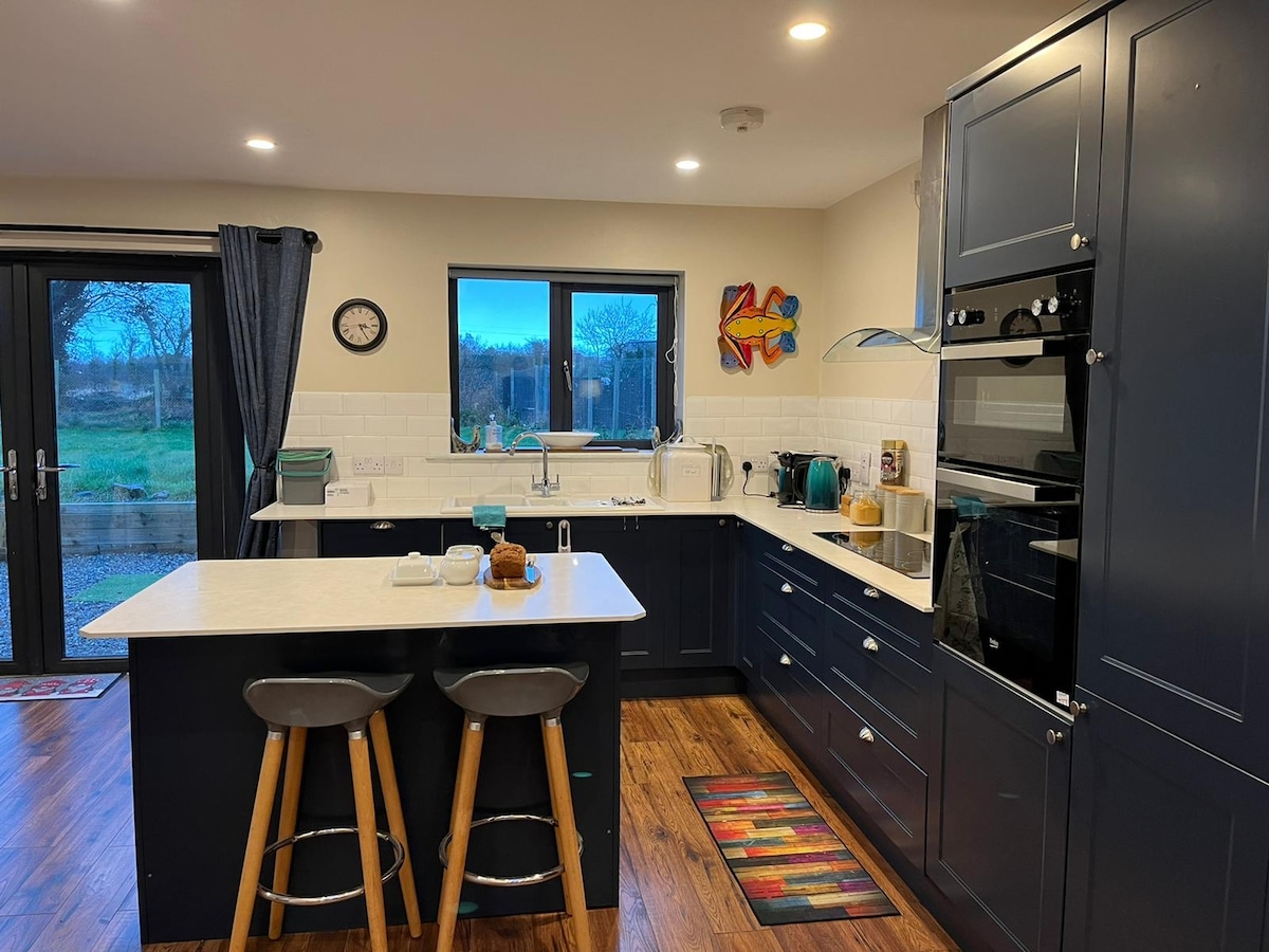 An open-plan kitchen showcases modern cabinetry in deep blue, complemented by light countertops. Bar stools are positioned at the island, creating a casual dining space. An assortment of kitchen appliances, including a kettle and coffee maker, are neatly arranged. Natural light enters through the large windows, illuminating the area.
