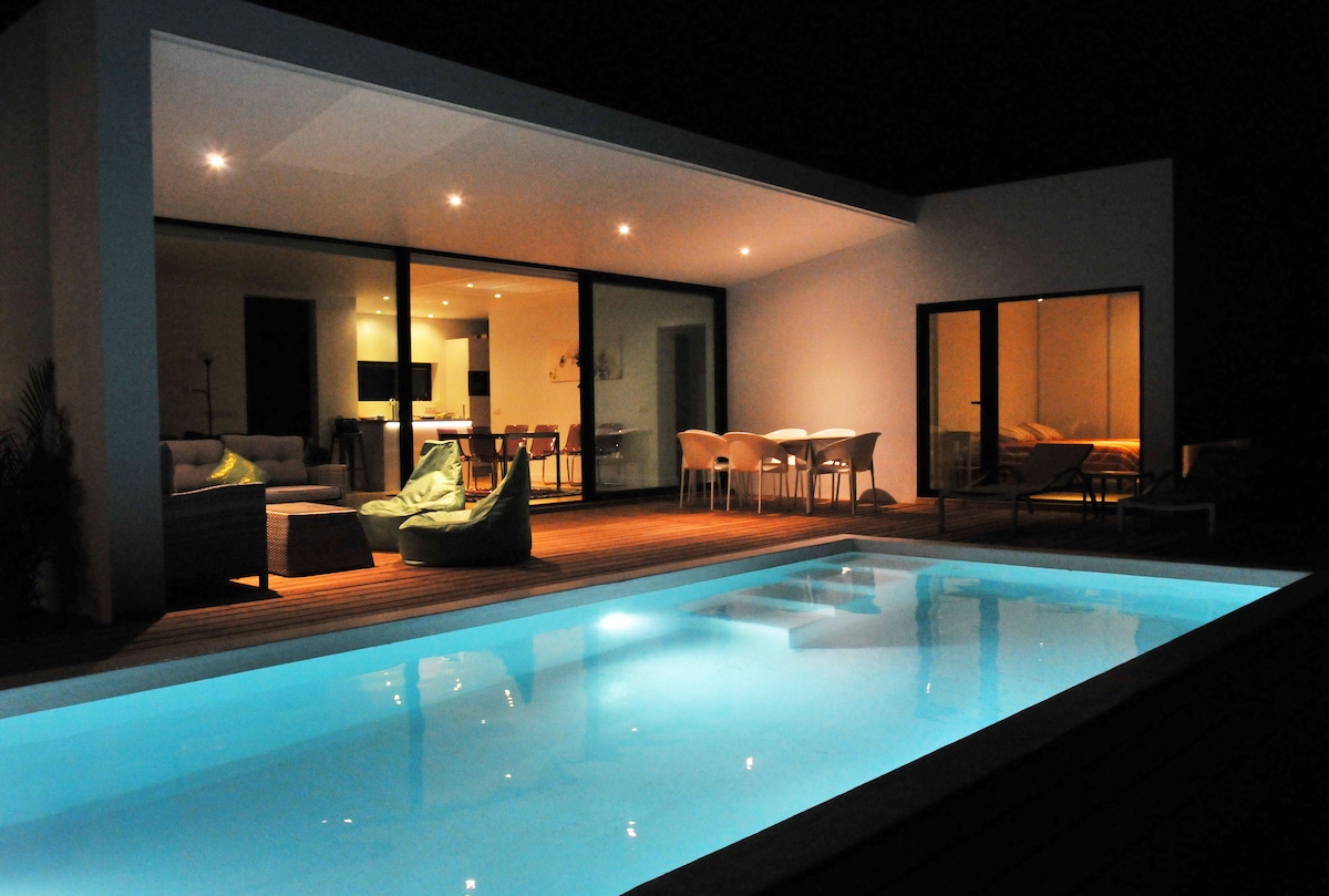 A modern villa at night features a serene, illuminated pool reflecting the soft glow of surrounding lights. The open living area showcases large glass doors that connect to an outdoor space, complemented by cozy seating and a dining area.
