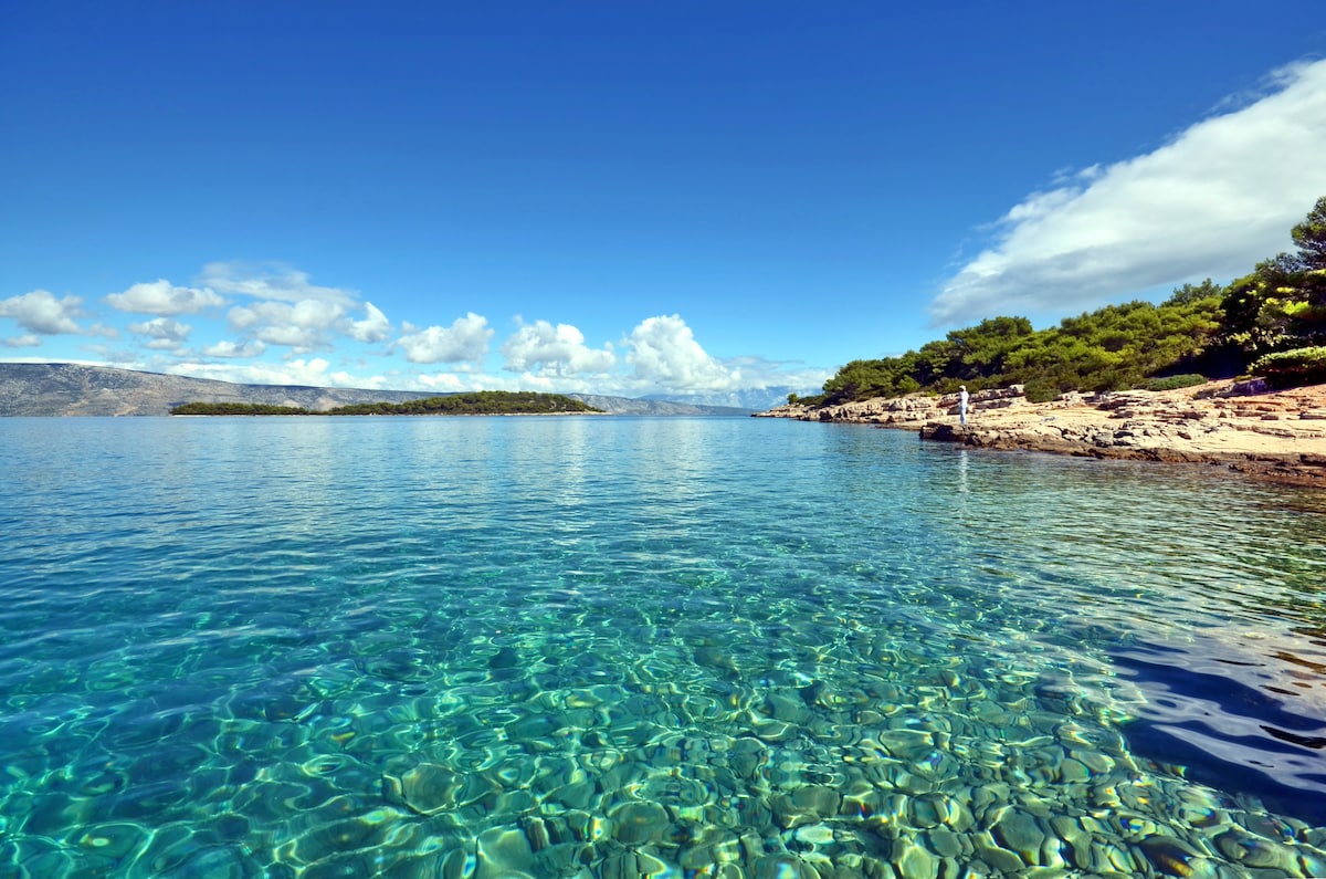 Clear turquoise waters reflect the blue sky, creating a serene vista. Sunlight glimmers on the water's surface, while rocky shorelines and scattered greenery frame the tranquil scene. The horizon offers a view of distant islands and soft clouds.