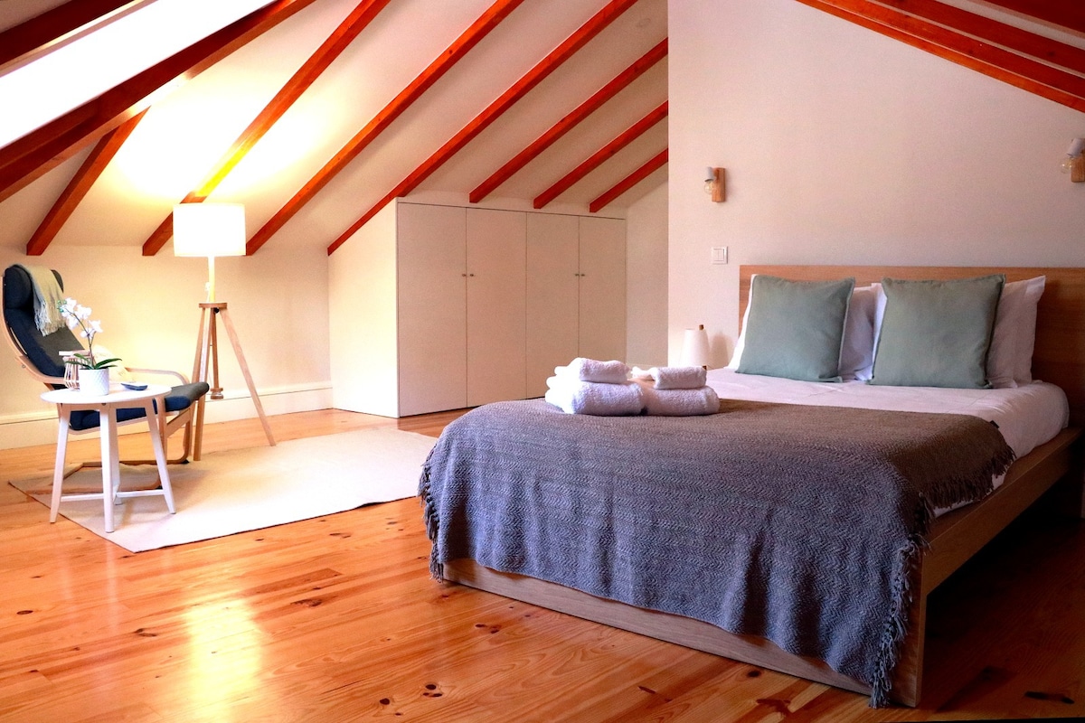 A spacious bedroom features a large bed with soft linens and decorative pillows. Natural wood flooring complements the warm tones of the sloped ceiling with exposed beams. A cozy armchair is positioned nearby, and a lamp provides gentle illumination, enhancing the inviting atmosphere.