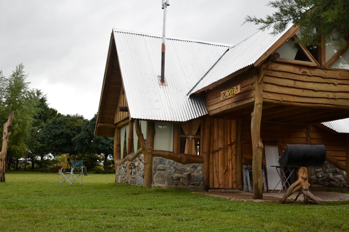 A rustic cabin constructed of wood and stone is featured, showcasing a sloped metal roof. The building is set within a grassy area, with a comfortable seating arrangement visible beside the cabin. Trees provide a natural backdrop, creating a serene outdoor environment.