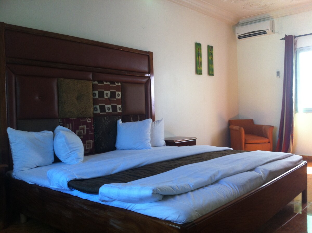 A spacious bedroom is furnished with a large bed adorned with white linens and decorative pillows. A cozy armchair sits nearby, and a window allows natural light to brighten the room. Air conditioning is present for comfort.