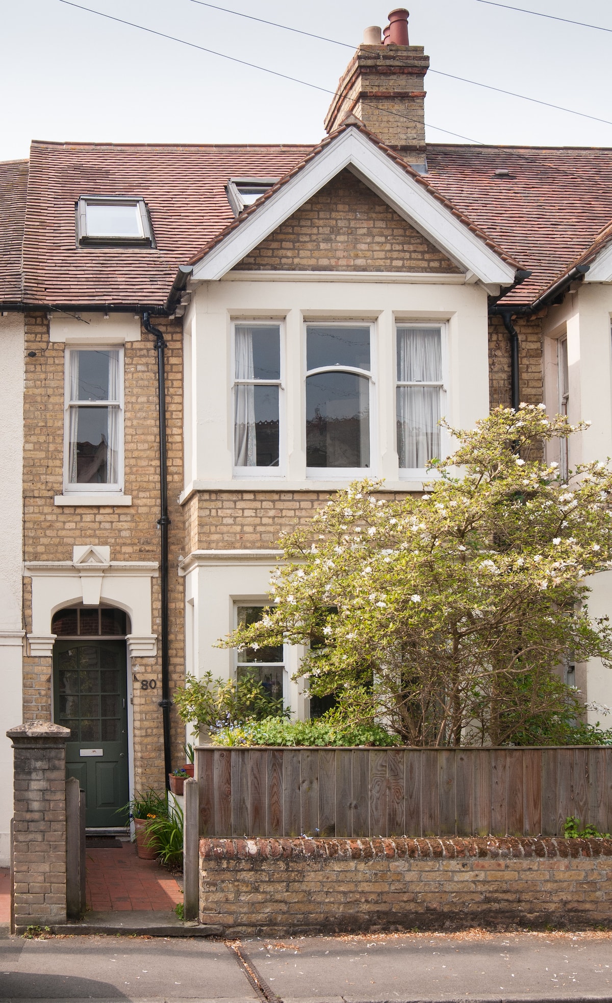 A Victorian terraced house is displayed, featuring a traditional facade with brickwork and a peaked roof. A low wooden fence surrounds a small garden with shrubs. Large windows allow natural light to illuminate the interior, enhancing the home's inviting exterior.