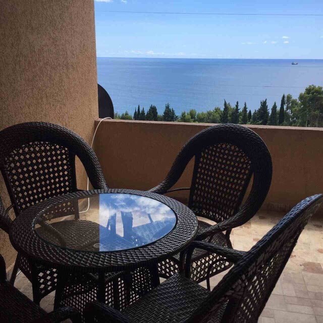 A balcony is presented featuring a round glass table surrounded by four wicker chairs. The expansive view of the sea is visible in the background, with blue skies and distant trees enhancing the tranquil setting.